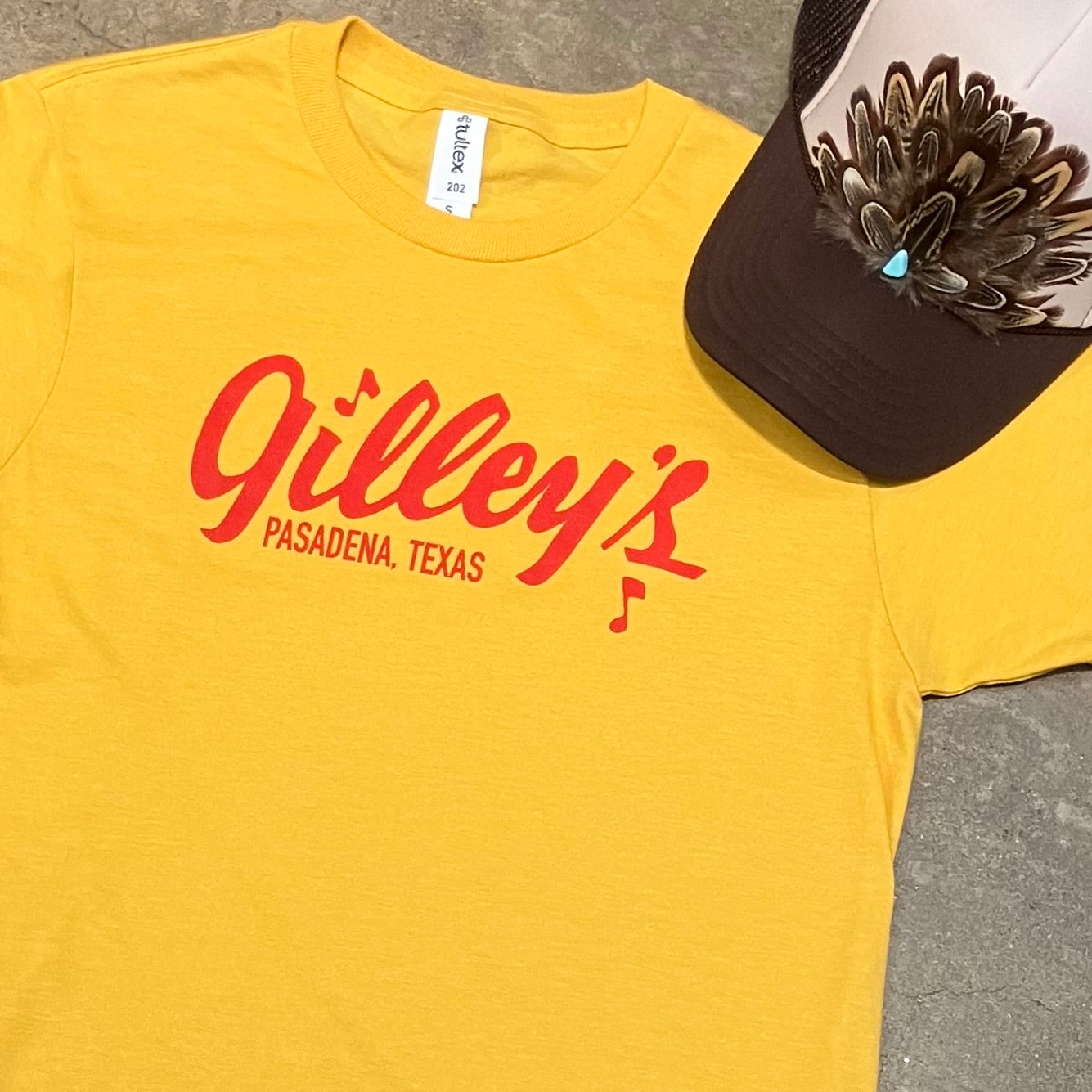 Heathered yellow tshirt with red Gilley’s Pasadena Texas logo screen printed on it. photo also features a trucker hat with feather hat band type embellishment.