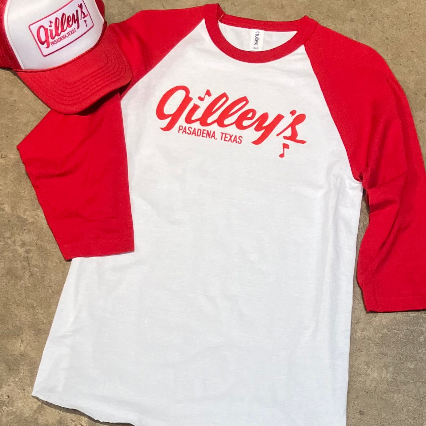 White and reg raglan baseball style tshirt with red Gilley's Pasadena Texas logo screen printed on it. Shown with a red mesh and white foam trucker hat featuring a Gilley's logo patch. 