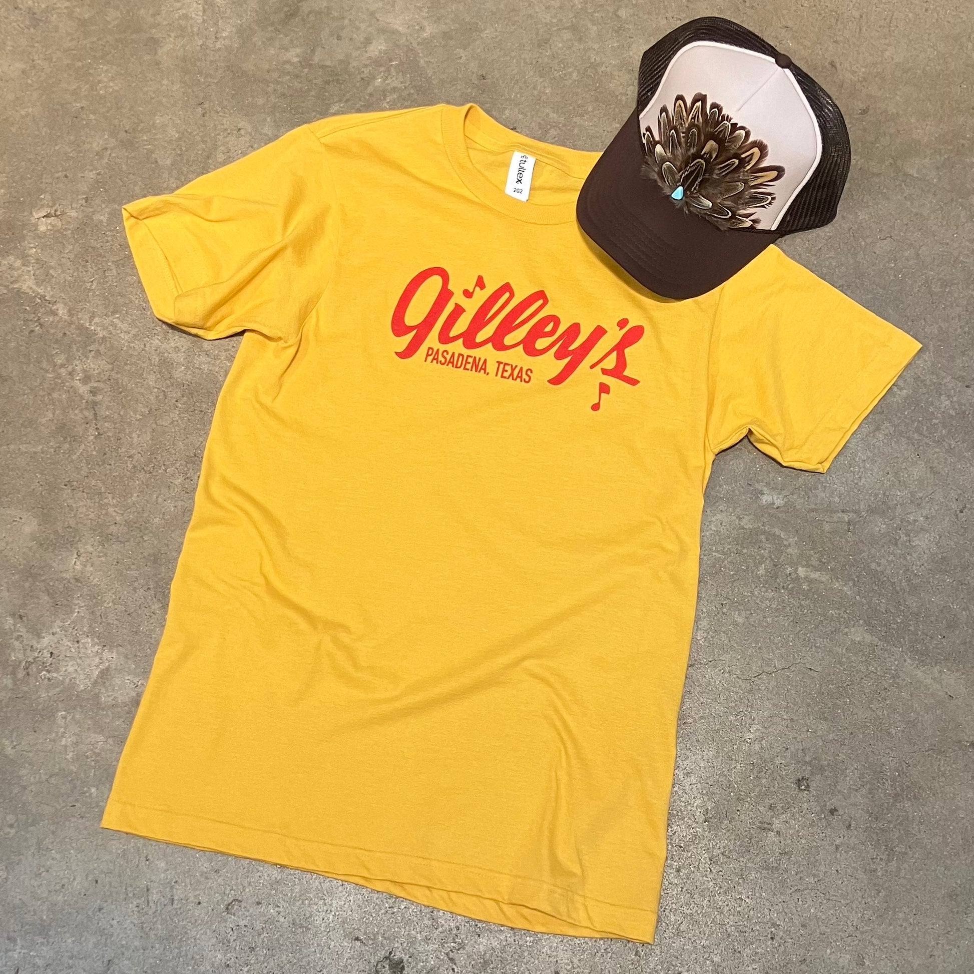 Heathered yellow tshirt with red Gilley’s Pasadena Texas logo screen printed on it. photo also features a trucker hat with feather hat band type embellishment.